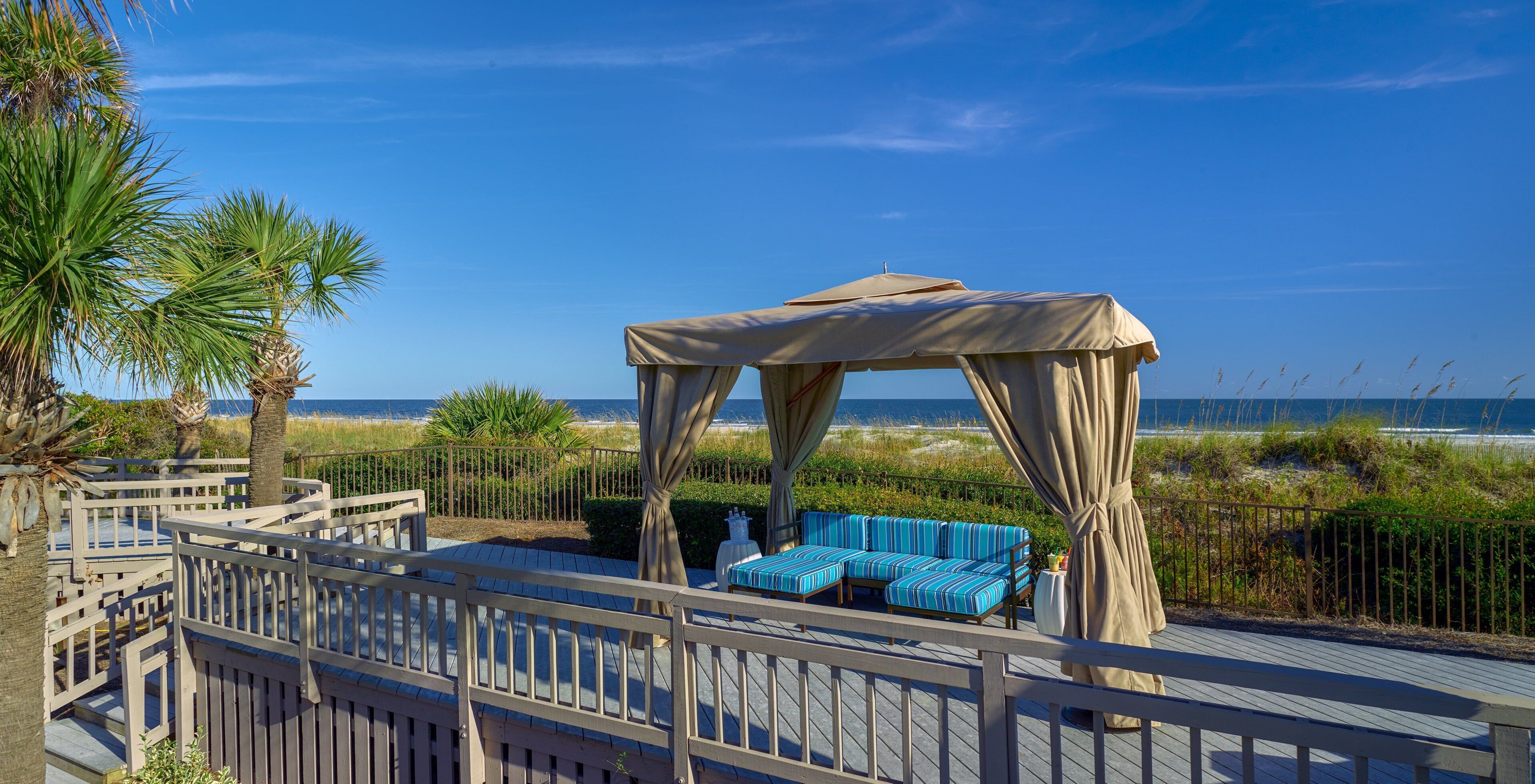 A wooden deck with a covered seating area, a view of the beach, and a palm tree.