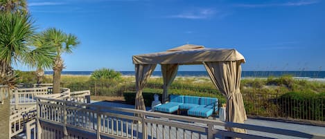 Una terraza de madera con zona de estar cubierta, vista a la playa y una palmera.