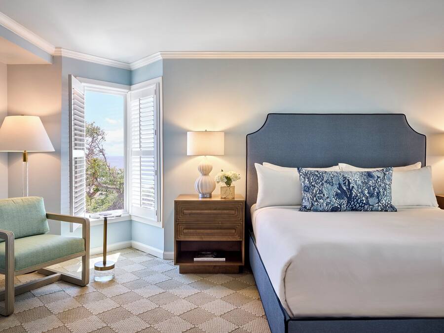 A bedroom with a bed, a chair, a nightstand with lamps, and a window with shutters.