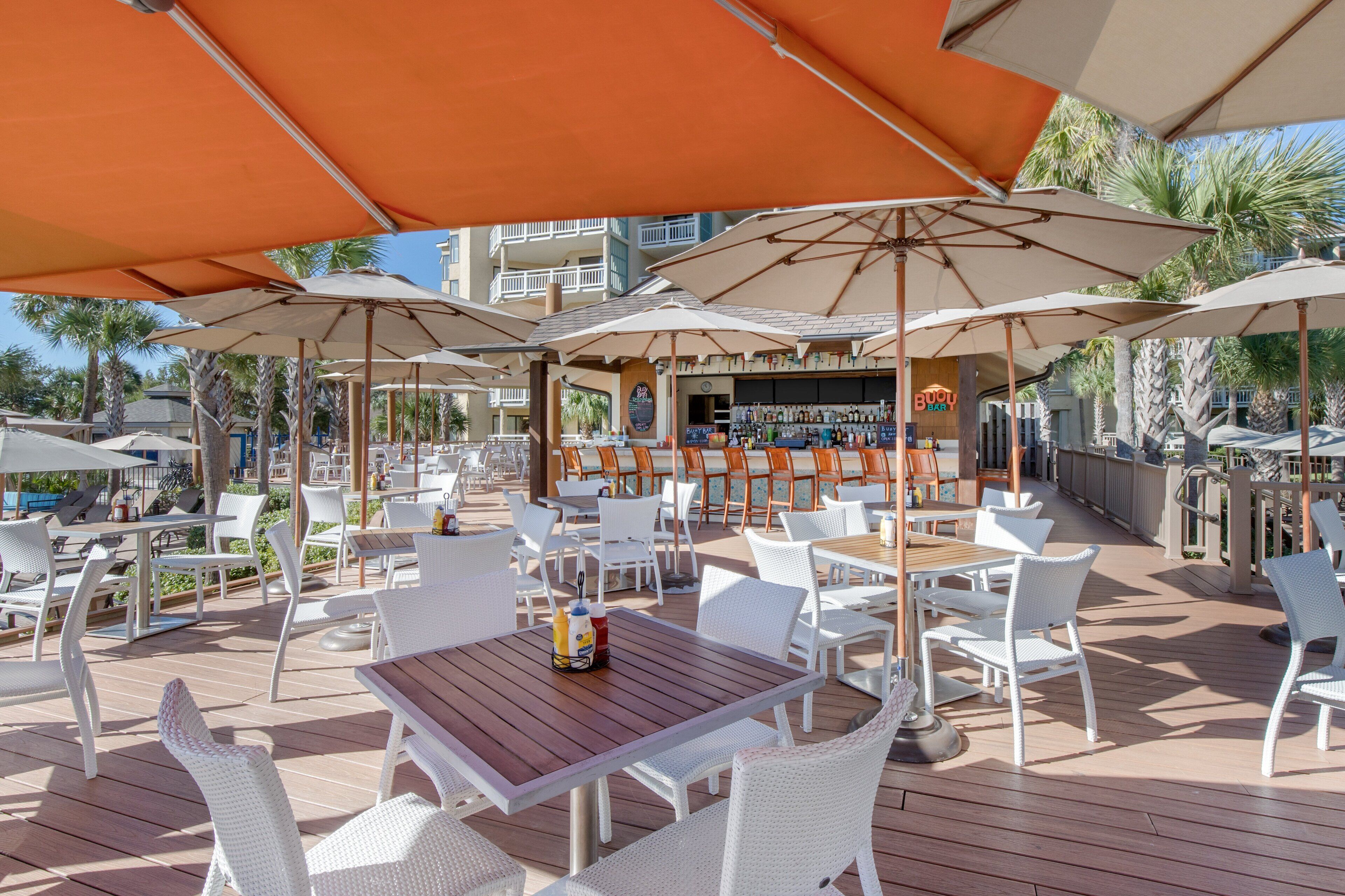 An outdoor patio with white wicker furniture, orange umbrellas, and a bar area with a 'BUDDY' sign.