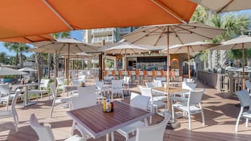 An outdoor patio with white wicker furniture, orange umbrellas, and a bar area with a 'BUDDY' sign.
