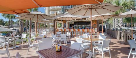 An outdoor patio with white wicker furniture, orange umbrellas, and a bar area with a 'BUDDY' sign.