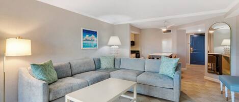 A modern living room with a grey sectional sofa, a white coffee table, and a floor lamp. There is a painting on the wall and a kitchen area in the background.