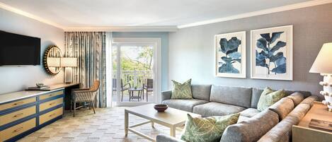 Una sala de estar moderna con sofá, mesa de centro y televisor sobre un mueble. Hay un balcón con vista a zonas verdes.