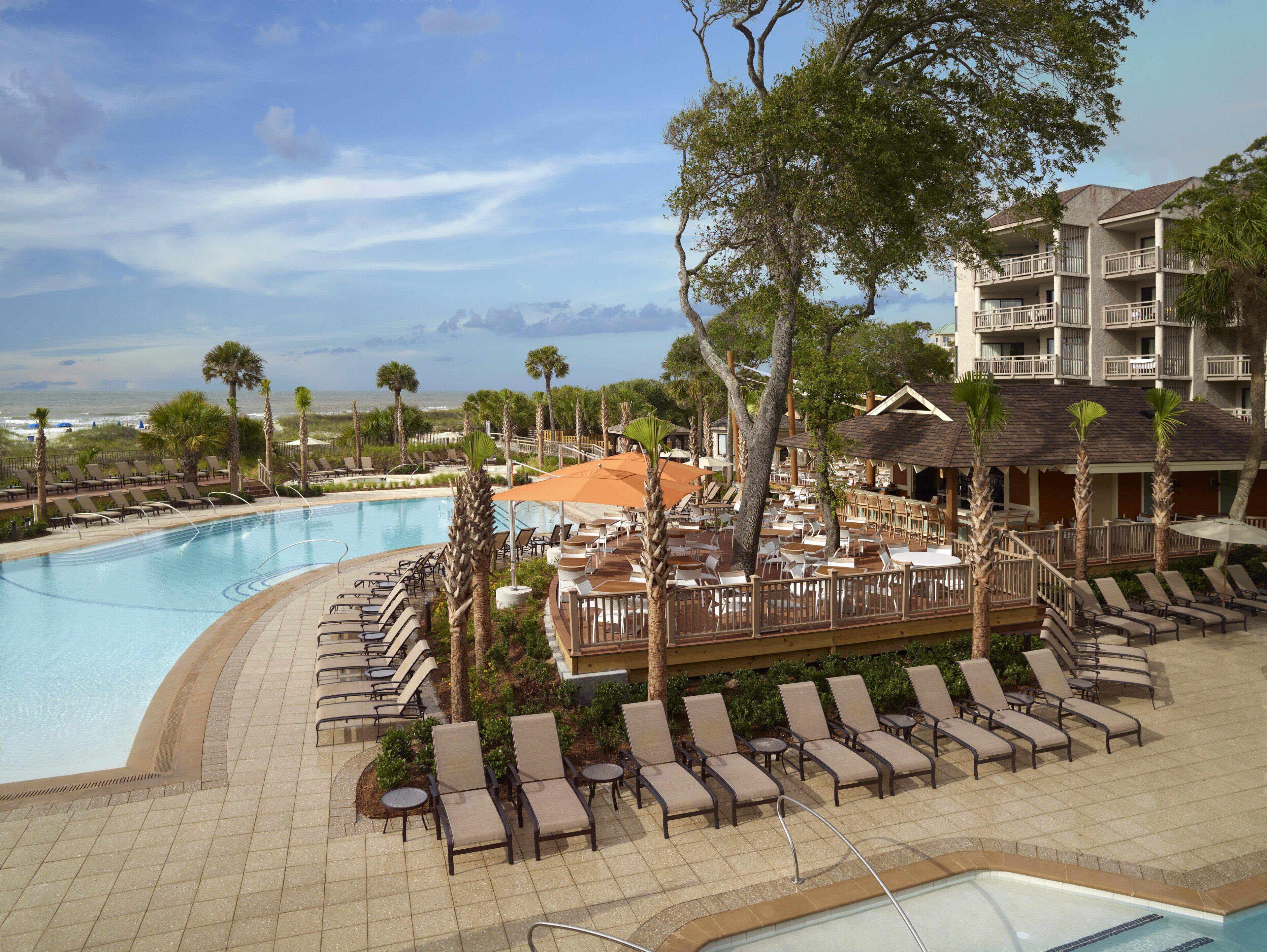 A resort pool area with lounge chairs and umbrellas, overlooking a beach and ocean.