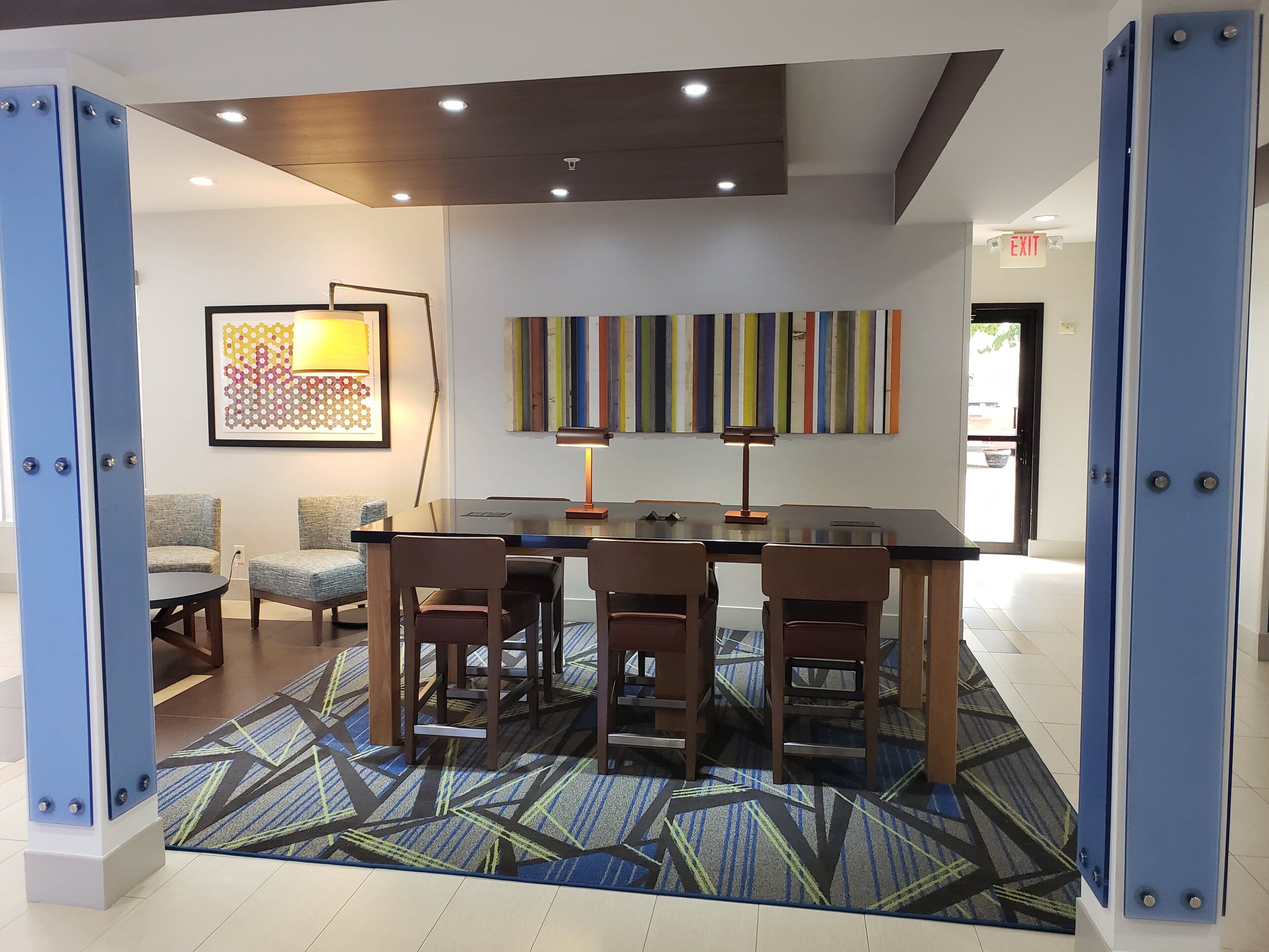 A modern hotel lobby with a conference table, chairs, a patterned rug, a wall art piece, and a colorful striped wall decoration.