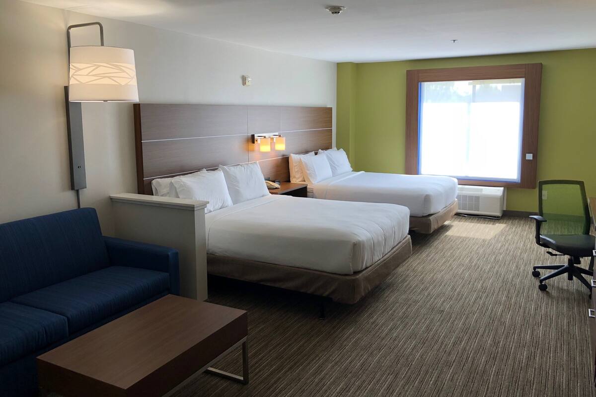 A hotel room with two beds, a blue sofa, a wooden coffee table, and a large window providing natural light.