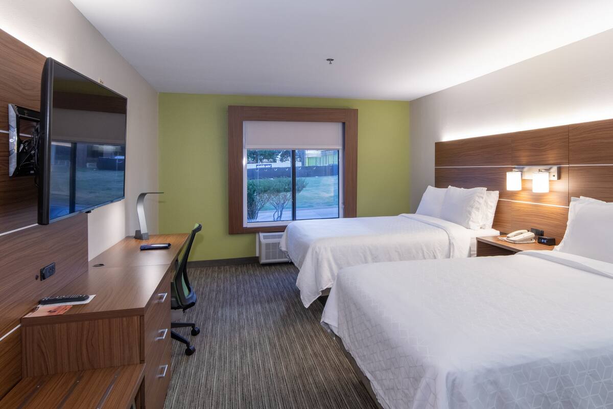 A hotel room with two beds, a desk, a flat-screen TV, a window with blinds, and a wall-mounted lamp.