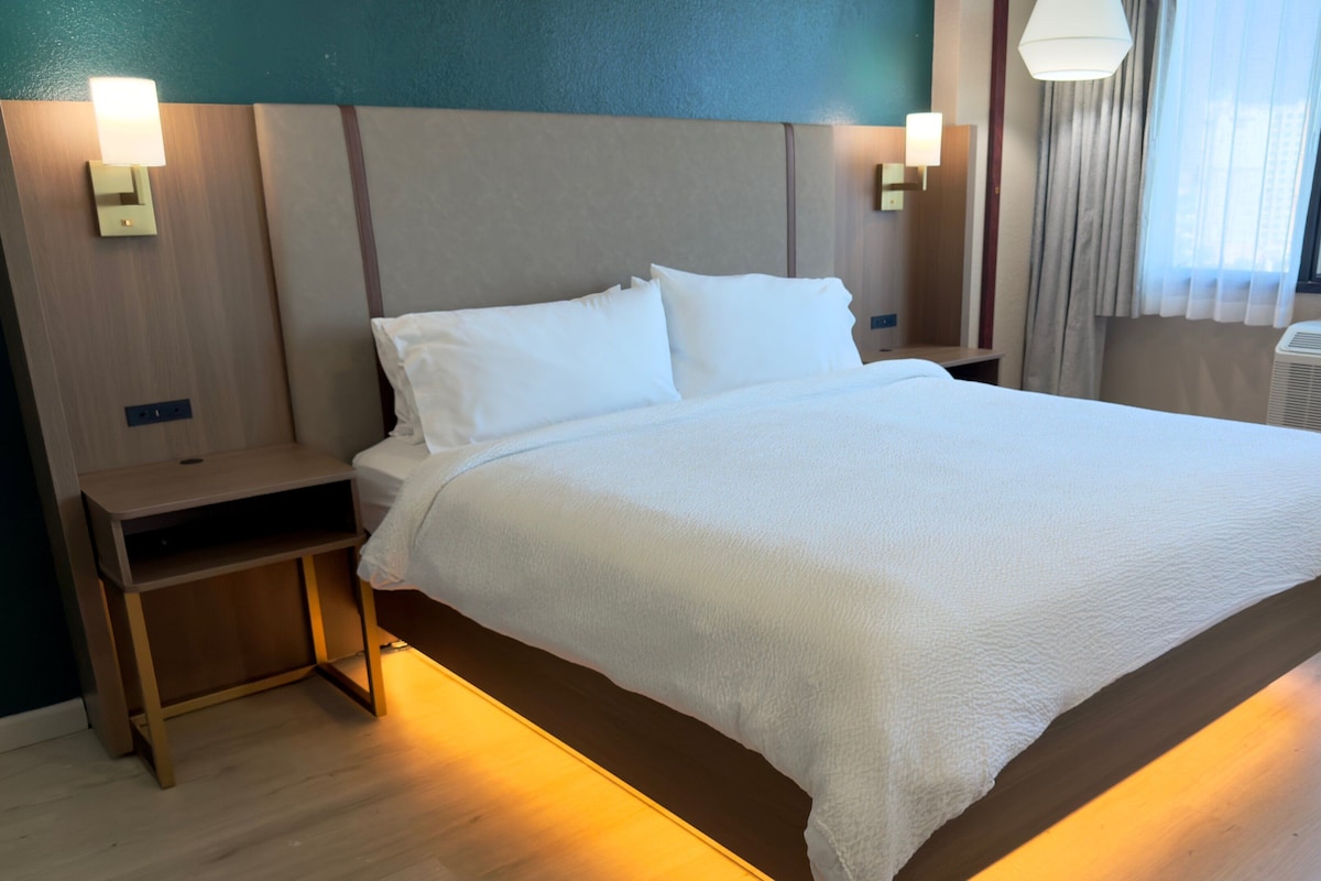 A hotel room with a large bed, bedside tables, and a view of the city through the window.