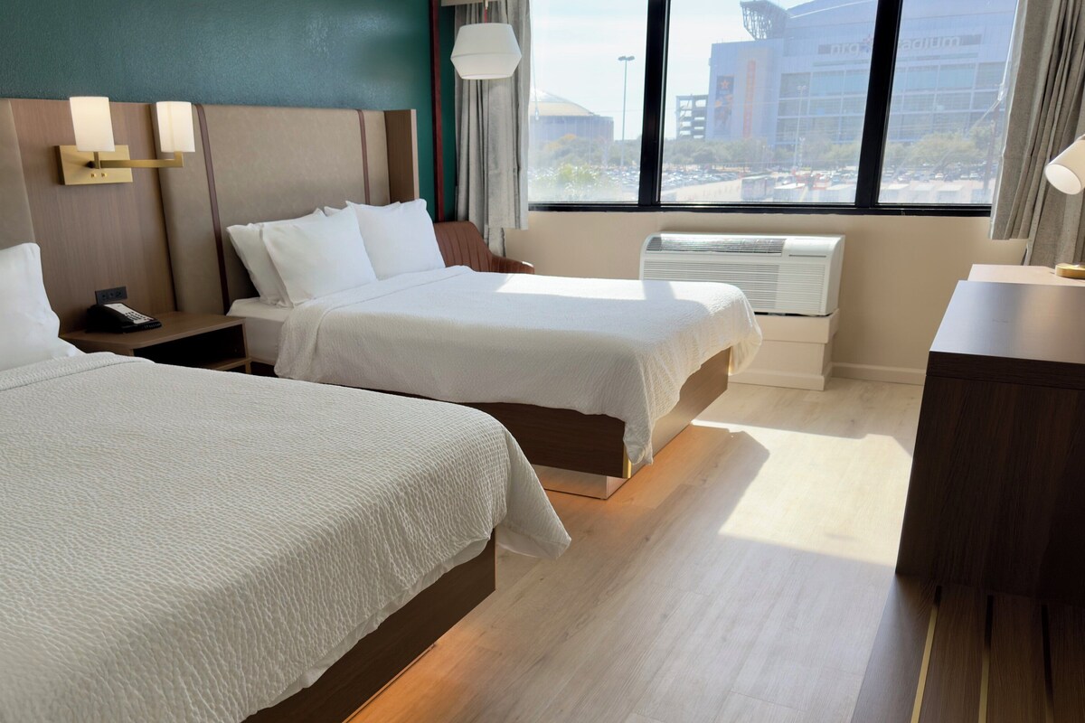 A hotel room with two beds, a bedside table with a phone, a lamp, and a view of buildings through the window.