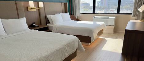 A hotel room with two beds, a bedside table with a phone, a lamp, and a view of buildings through the window.