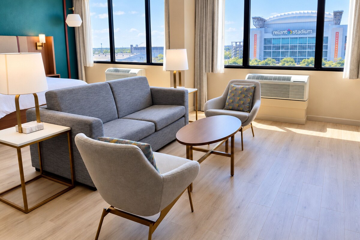 Quarto de hotel com sofá cinzento, poltronas, uma mesa redonda pequena e vista para um estádio.