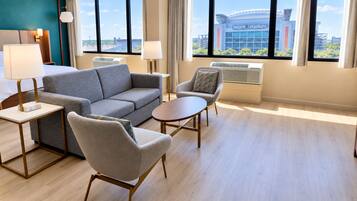 A hotel room with a grey sofa, armchairs, a small round table, and a view of a stadium.