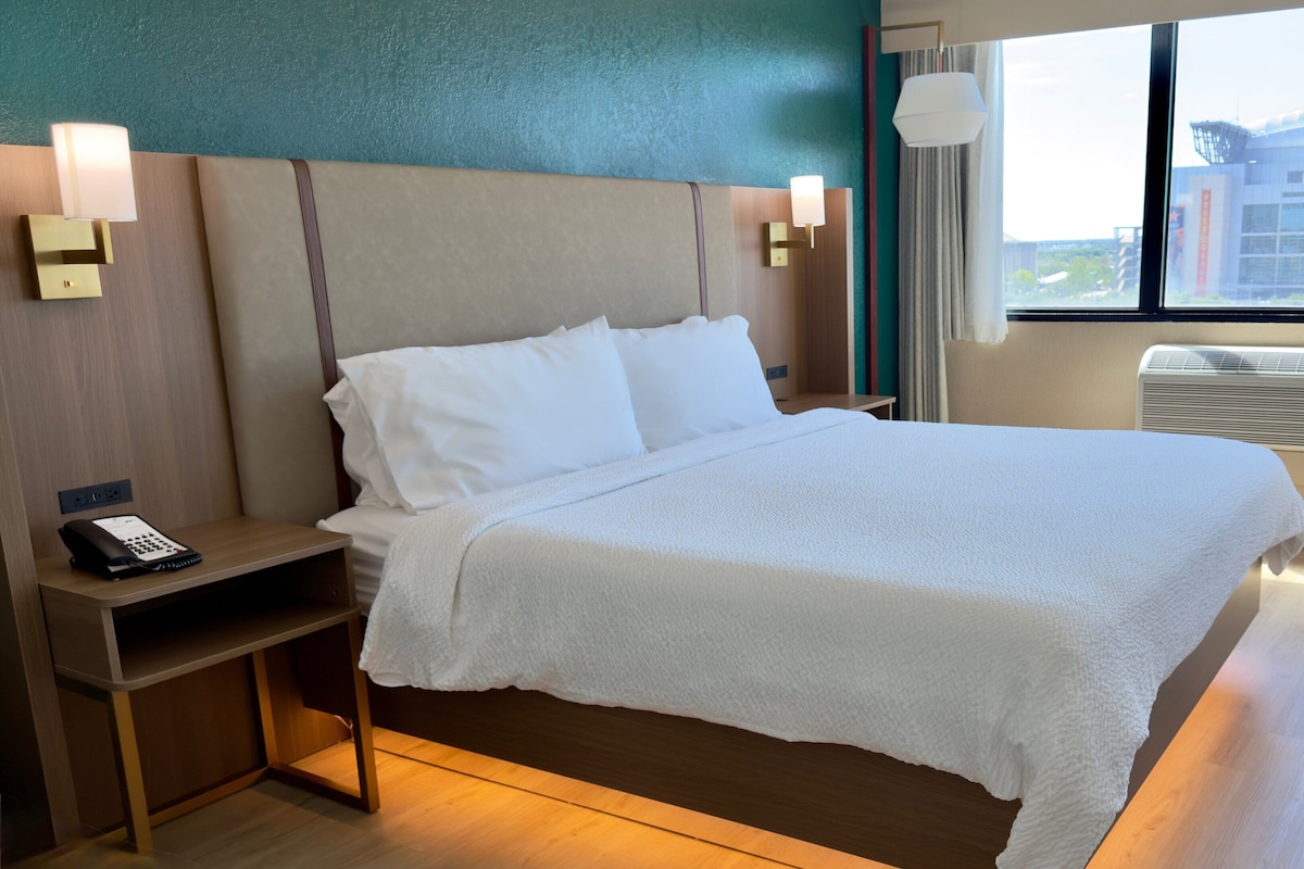 A hotel room with a bed, bedside table, lamp, and window with a view of a building.