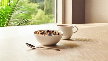 A bowl of cereal with a spoon on a wooden table by a window with a view of greenery.