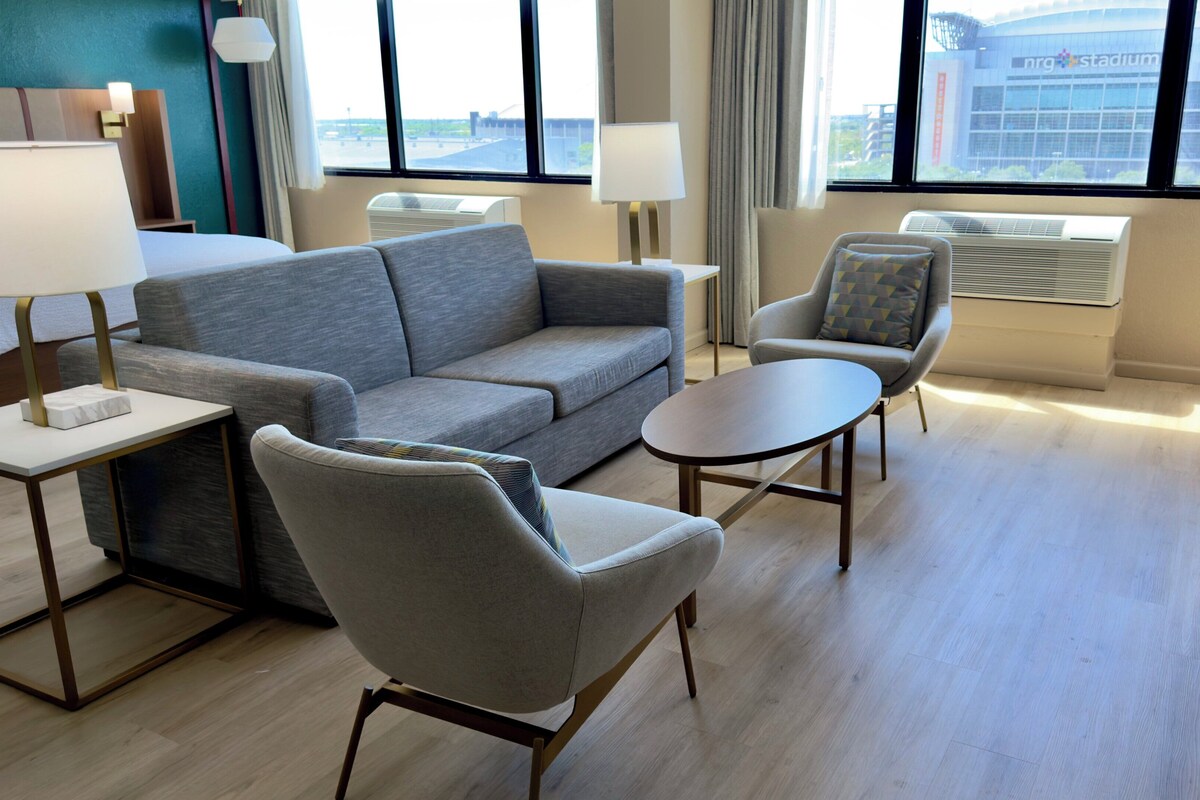 A modern hotel room with a grey sofa, a round table, two armchairs, and a view of a stadium through large windows.