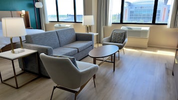Una habitación de hotel moderna con un sofá gris, una mesa redonda, dos sillones y vistas a un estadio a través de amplios ventanales.