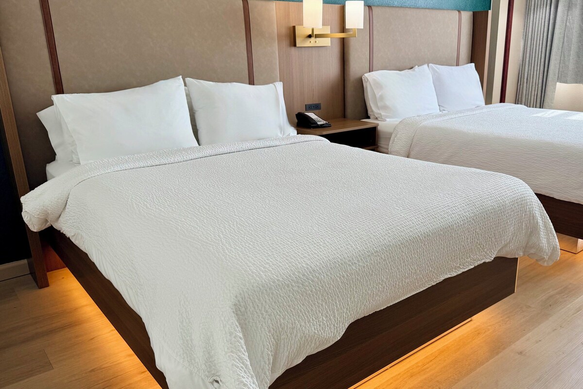 Two beds with white linens, a bedside table with a phone, and a wall-mounted light fixture.