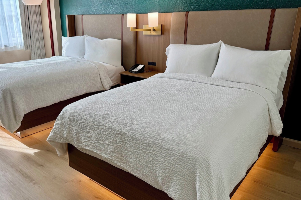 A hotel room with two beds, a bedside table with a phone, and a wall-mounted light fixture.