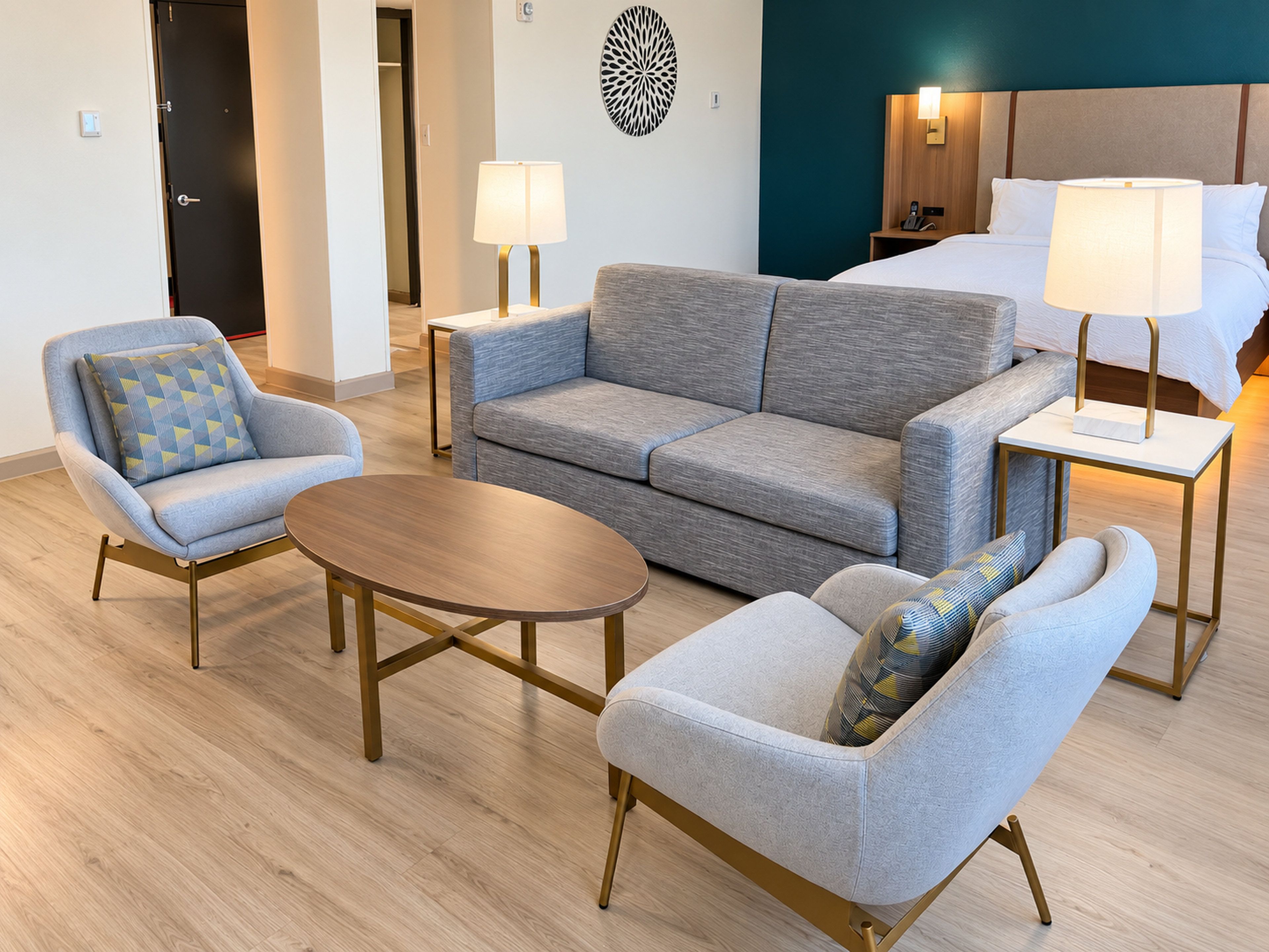 Una habitación de hotel moderna con un sofá gris, una mesita redonda y dos sillones. Se observa una cama con edredón blanco y dos lámparas de noche.