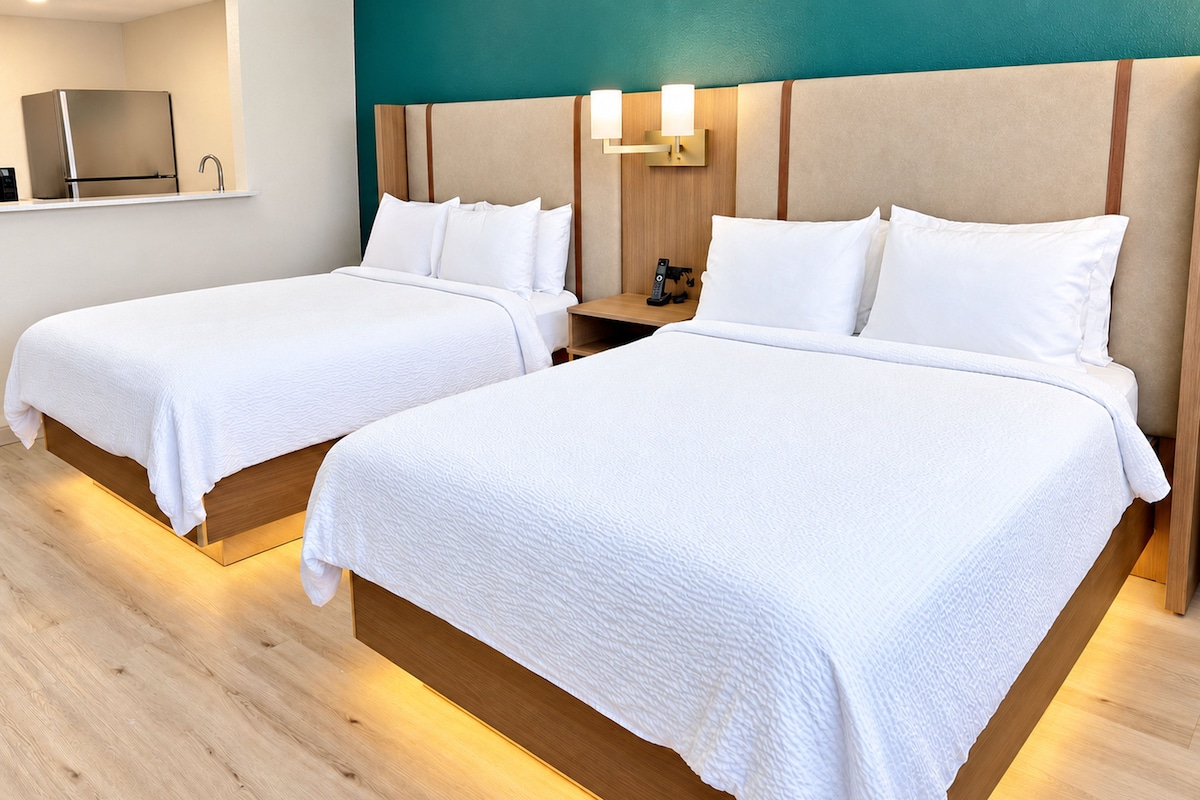 Two single beds with wooden headboards and white bedding, a nightstand with a lamp, and a television on a shelf above the beds.