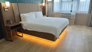 A hotel room with a bed, bedside table, lamp, and air conditioning unit.