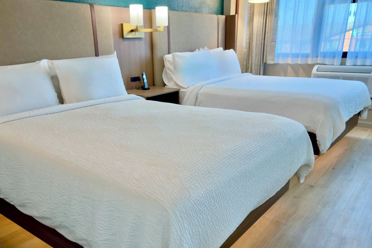 A hotel room with two beds, a nightstand with a lamp, and a window with curtains.