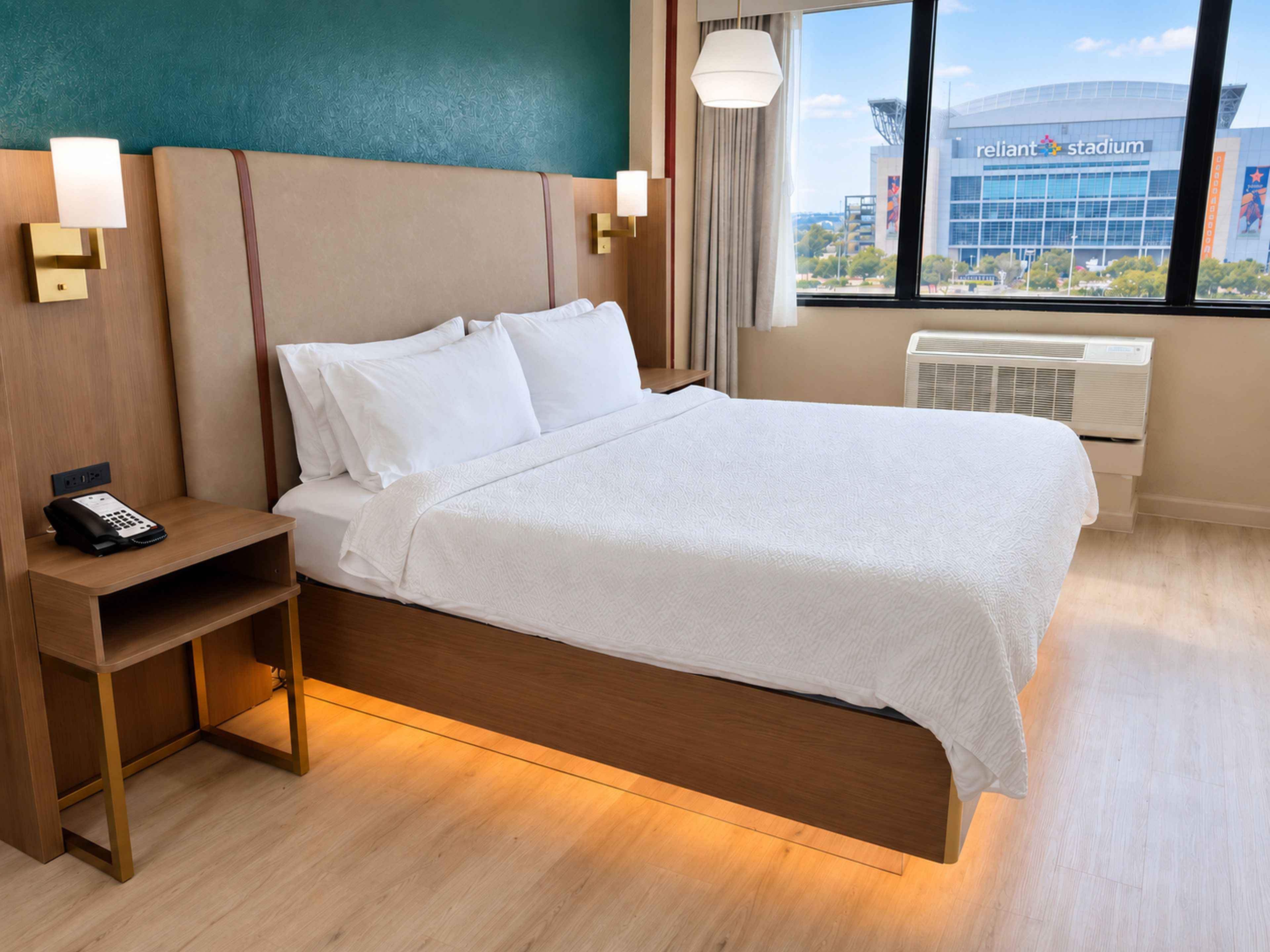 A hotel room with a bed, bedside table, lamp, and a view of a stadium.
