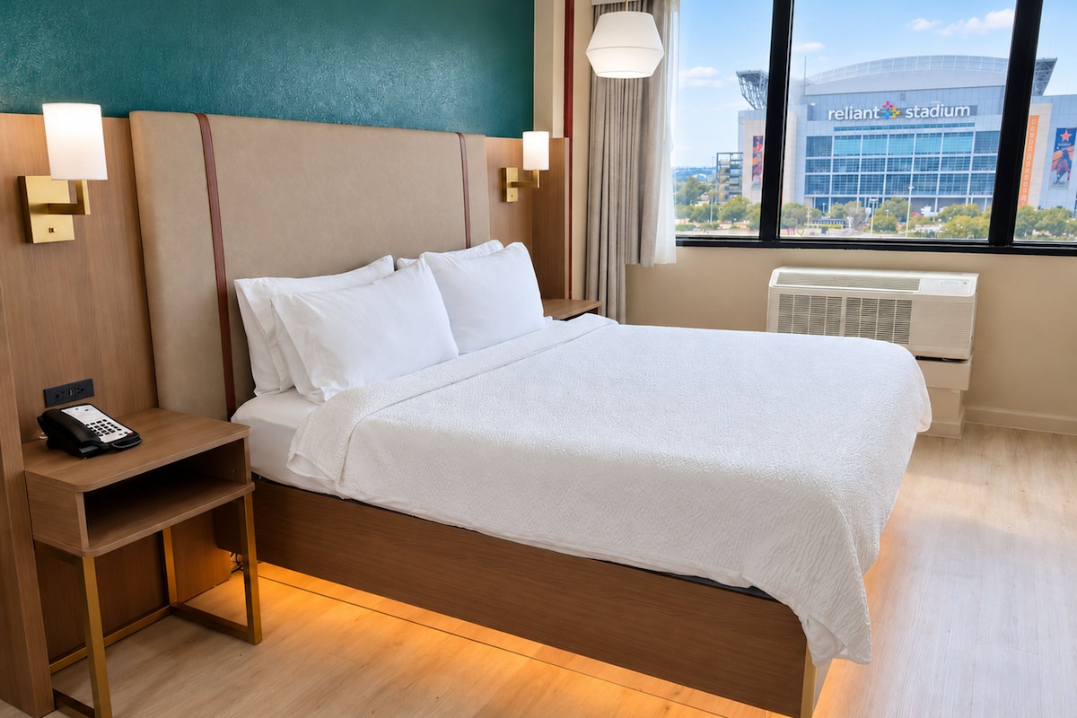 A hotel room with a bed, bedside table, lamp, and a view of a stadium.
