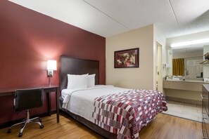 Une chambre d’hôtel avec un lit, un bureau, une chaise, une table de chevet, un tableau et une salle de bain visible à travers une porte ouverte.