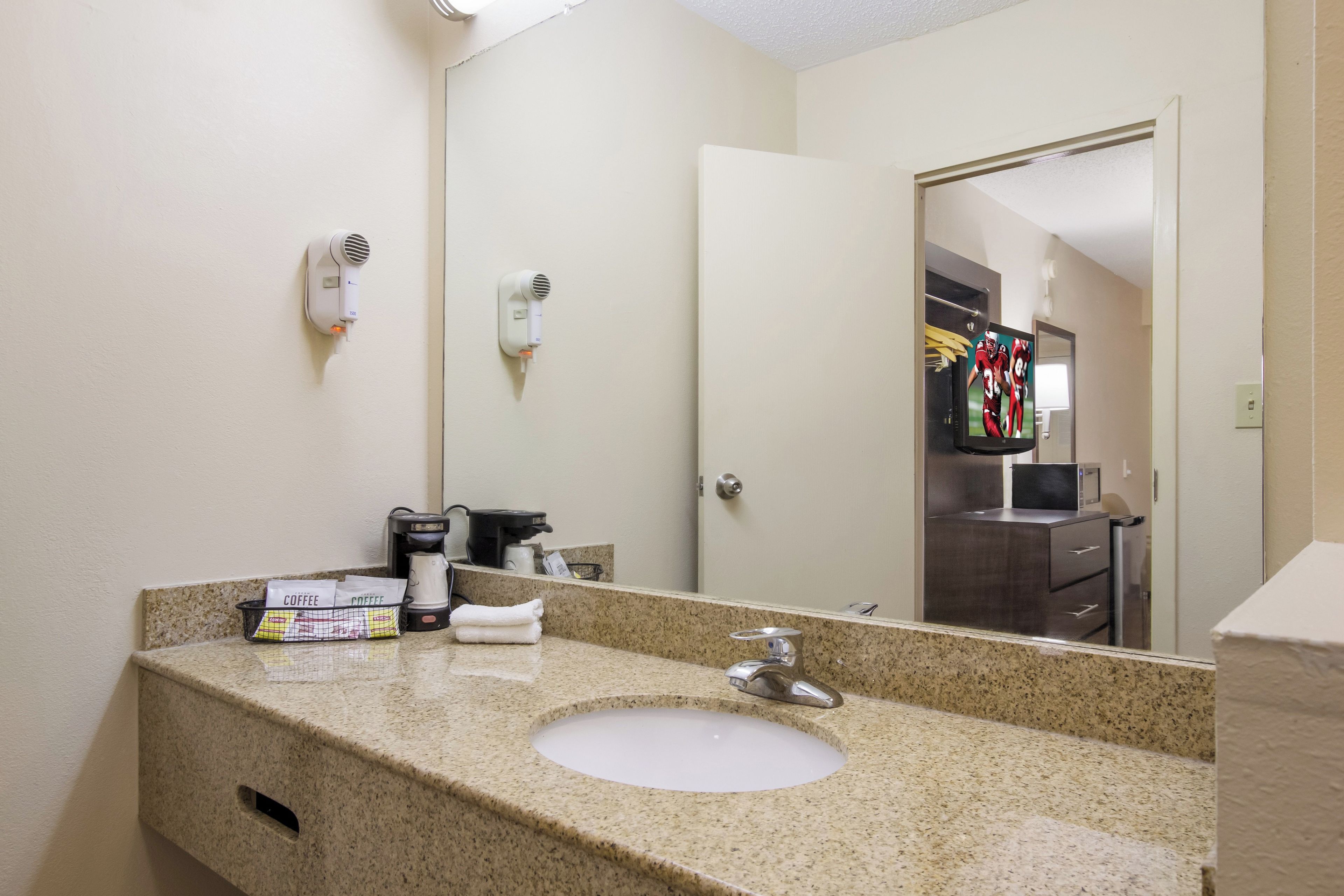 A hotel bathroom with a granite countertop, sink, and a mirror. There are hair dryers mounted on the wall, a coffee maker, and a towel rack.