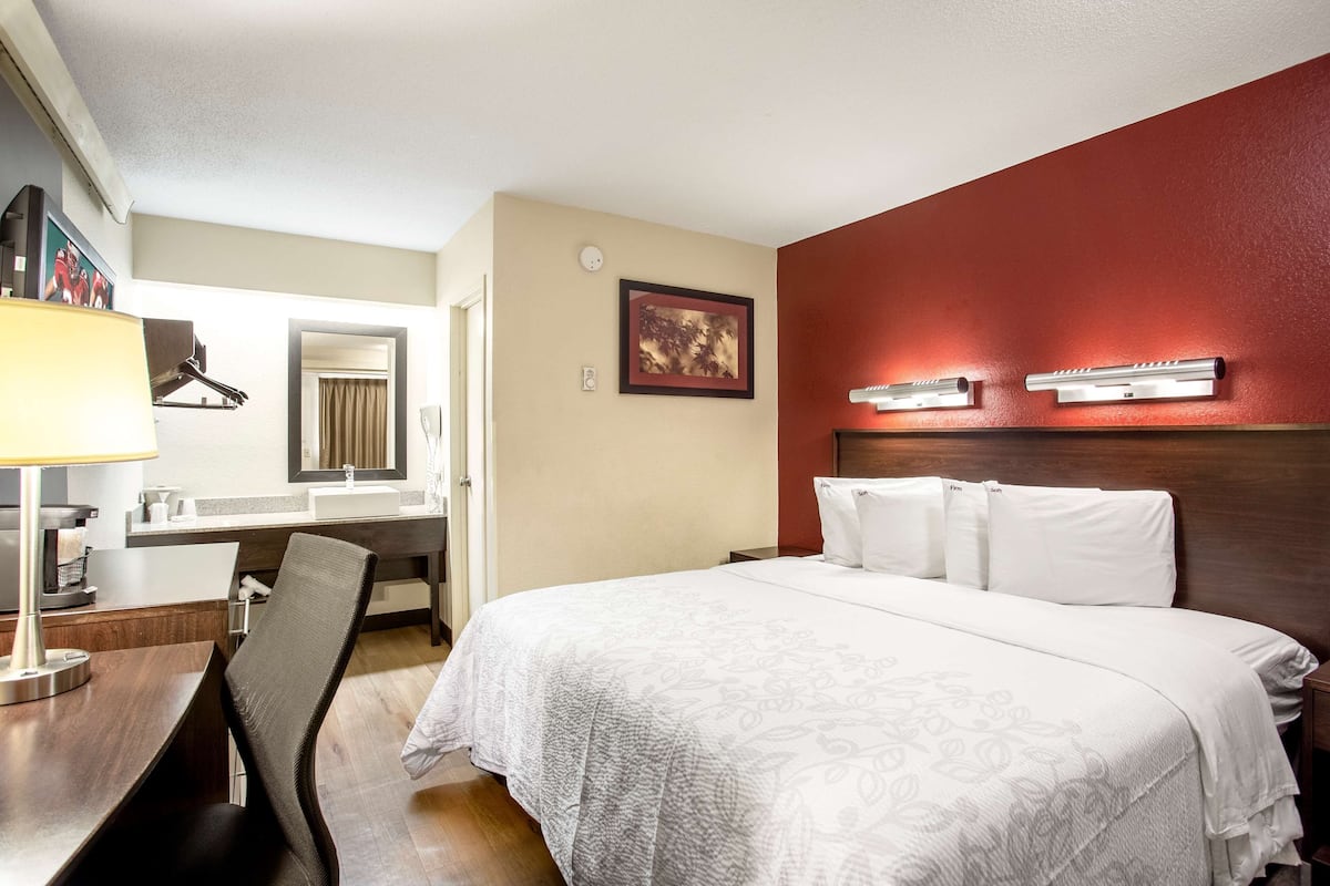 A hotel room with a bed, a desk, a chair, a desk lamp, a mirror, a painting, and a red accent wall.