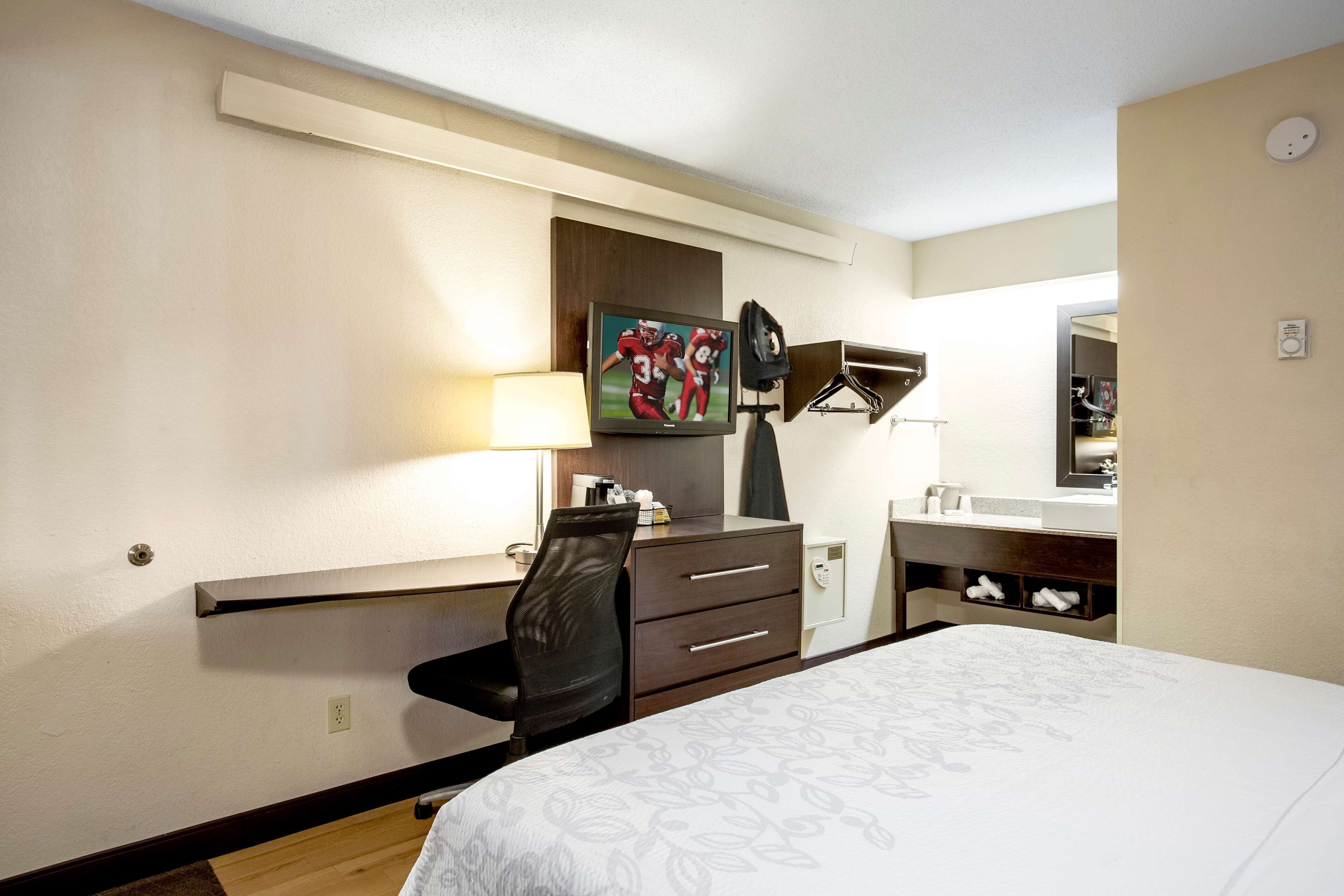 Una habitación de hotel con cama, escritorio con silla, televisor, espejo y perchero en la pared.