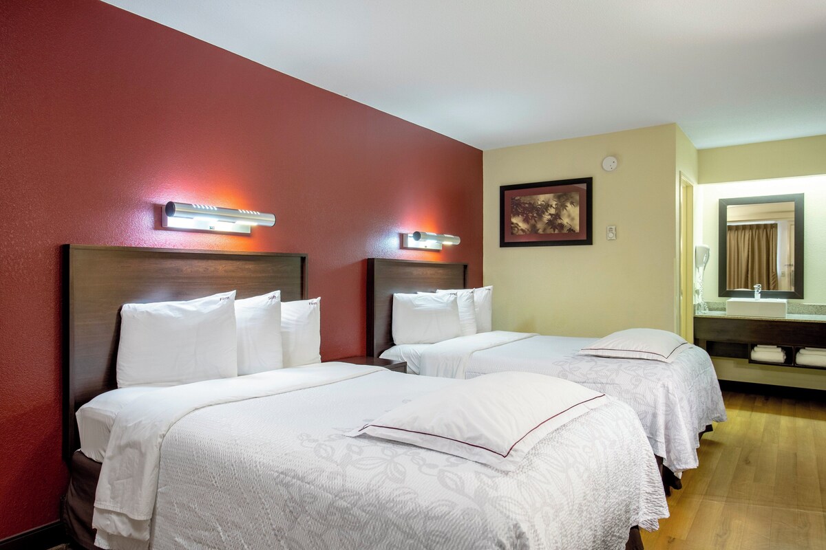 A hotel room with two beds, a wooden headboard, a wall-mounted light fixture, a framed picture, and a bathroom with a mirror and sink.