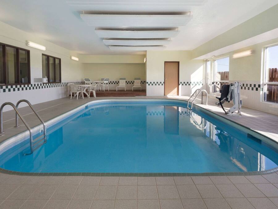 Una piscina interior con suelo de baldosas, una escalera de piscina y una zona de estar con sillas y una mesa.