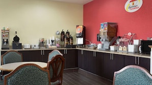 A breakfast area with a red wall, a counter with various food items, and seating with patterned chairs. - Super 8 by Wyndham Lees Summit (Lee's Summit)