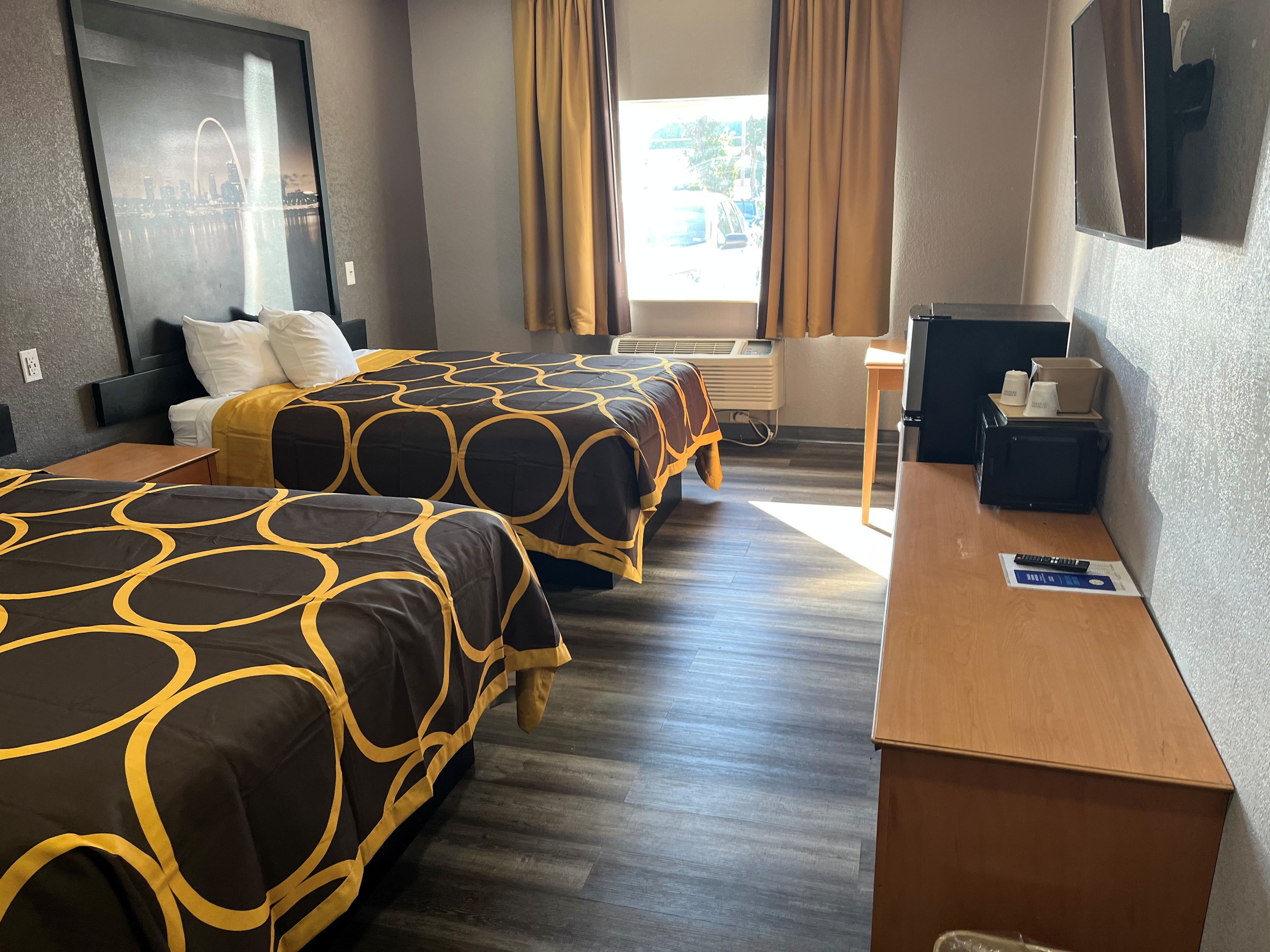 Ein Hotelzimmer mit zwei Betten, einem Schreibtisch, einem Stuhl, einem Flachbildfernseher und einem Fenster mit Vorhängen.