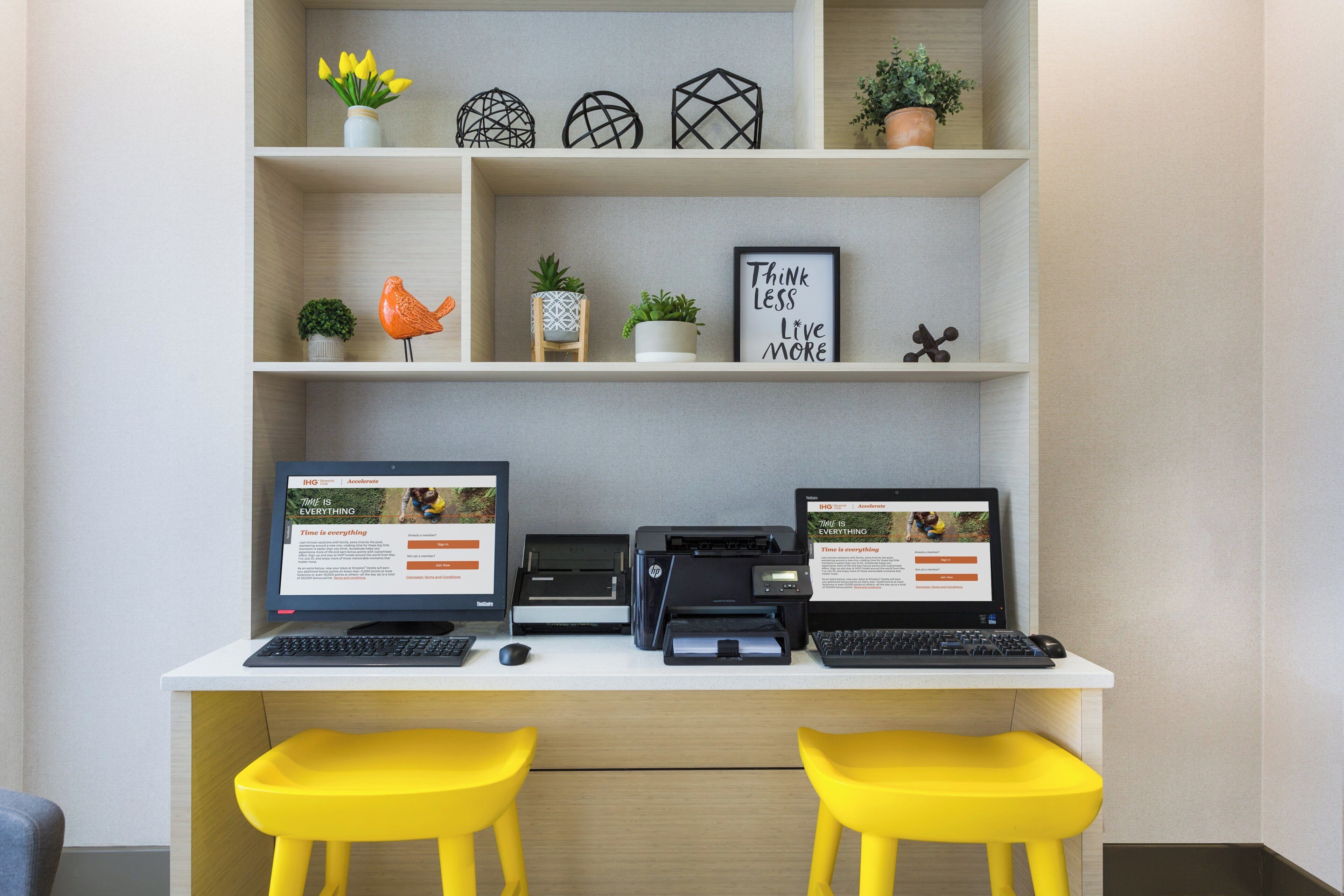 Ein Arbeitsplatz mit zwei Computern, einem Drucker und einem Schreibtisch mit gelben Hocker.