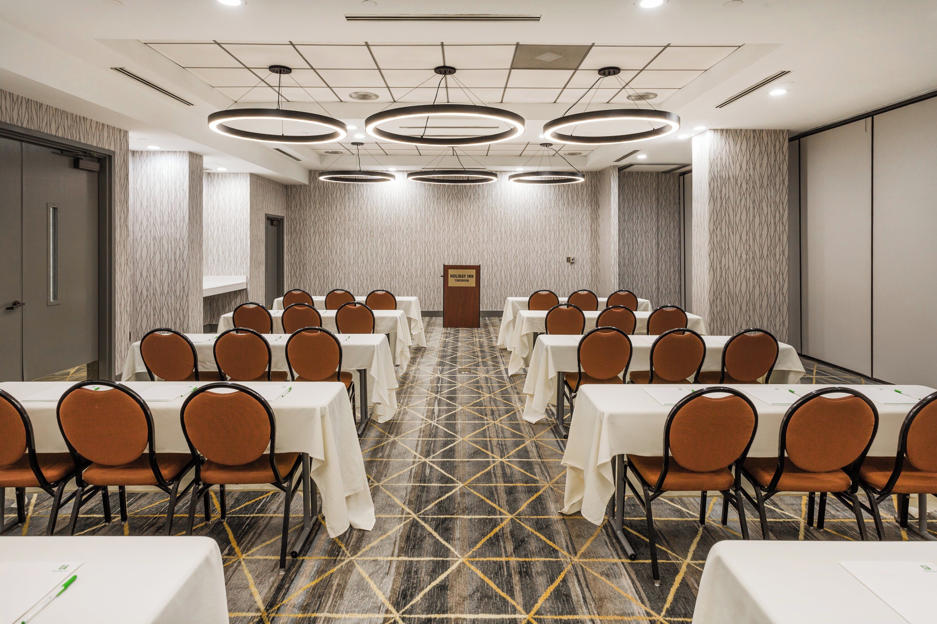 Un salón de conferencias con varias mesas y sillas, una alfombra estampada y una puerta con la indicación 'Holiday Inn TIMONUM'.