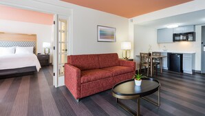 A hotel room with a bed, a red sofa, a small dining table with chairs, a kitchen area with a microwave and refrigerator, and a door leading to another room.