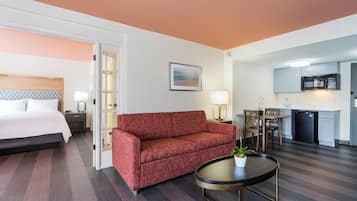 Habitación de hotel con cama, sofá rojo, una mesita con una planta, zona de cocina con microondas y una mesa de comedor con sillas.