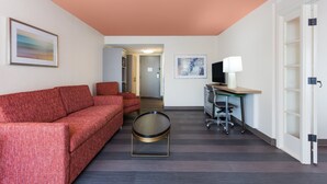 Une chambre d’hôtel moderne avec un canapé rouge, une table basse ronde, un bureau avec un ordinateur et une lampe, et un téléviseur posé sur un meuble.
