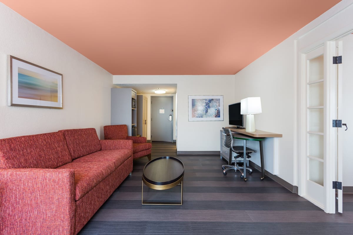 A modern hotel room with a red sofa, a round coffee table, a desk with a computer and a lamp, and a TV on a stand.