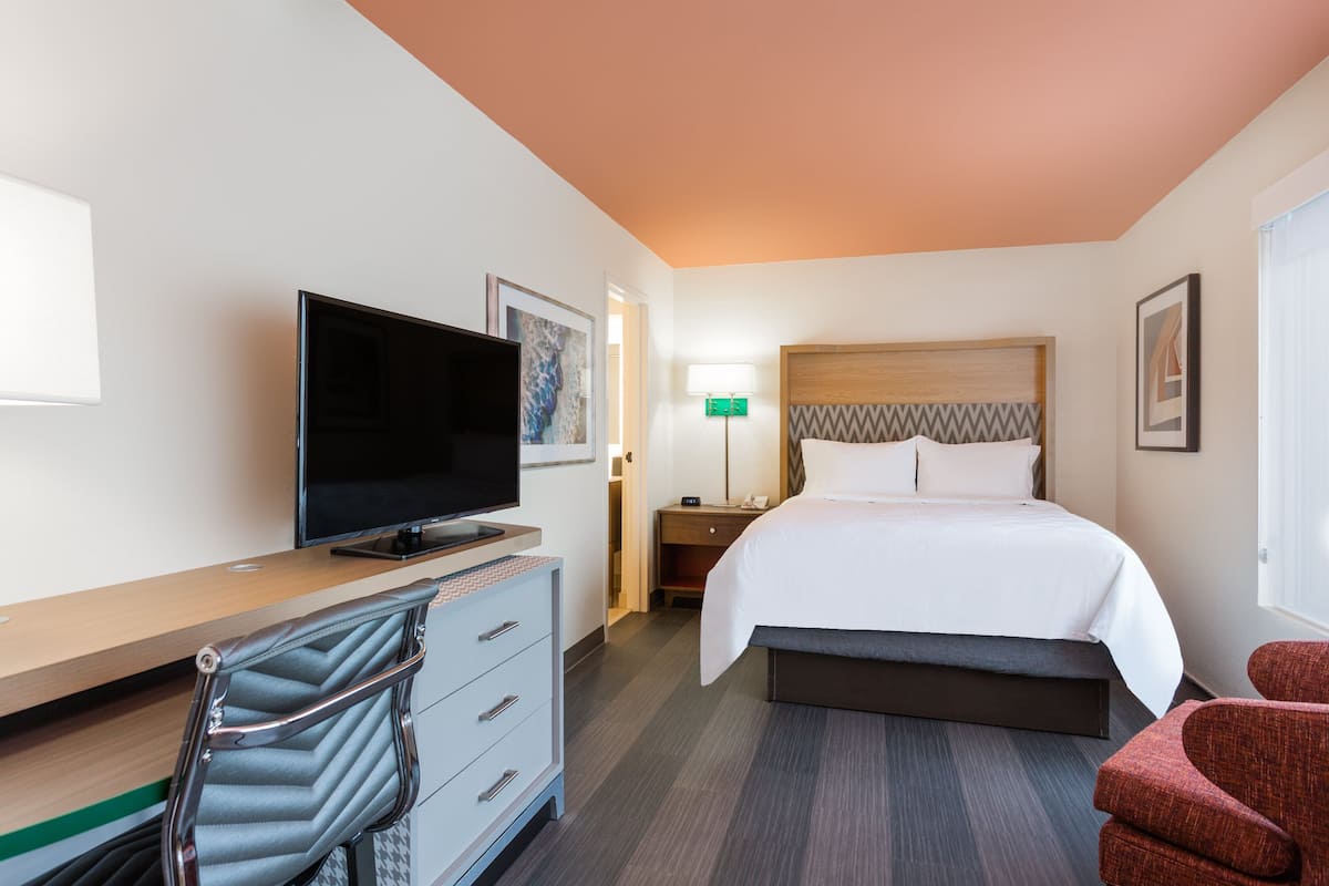 A hotel room with a bed, a TV, a desk with a chair, a nightstand, and a lamp.