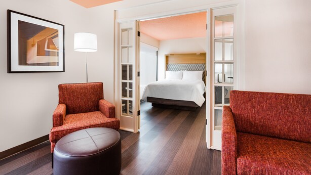 Une chambre d’hôtel avec un lit, un fauteuil rouge, un pouf rond et un tableau encadré au mur.