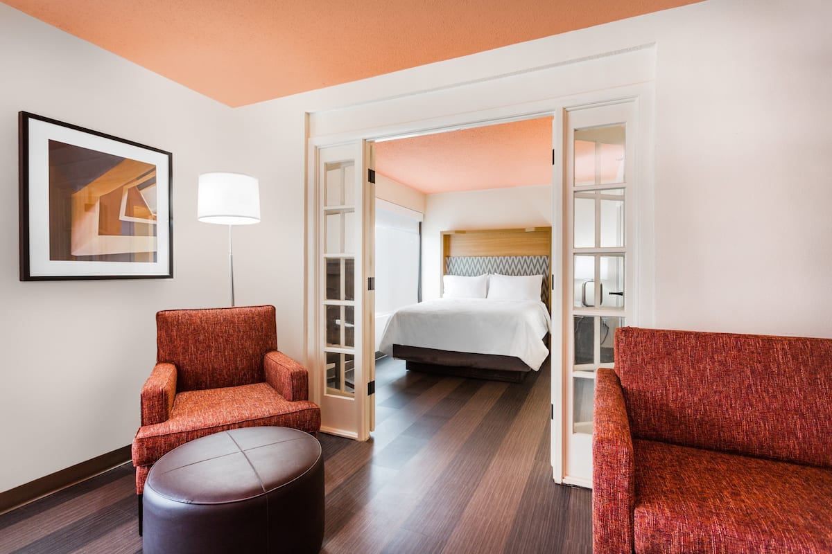 A hotel room with a bed, a red armchair, a round ottoman, and a framed picture on the wall.