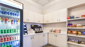 A small kitchenette with a refrigerator stocked with beverages, a coffee machine, and shelves with snacks.