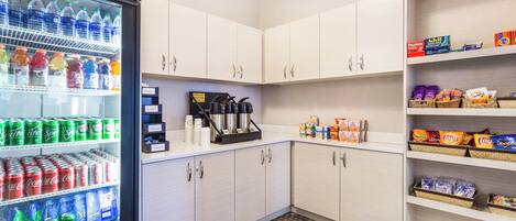 A small kitchenette with a refrigerator stocked with beverages, a coffee machine, and shelves with snacks.