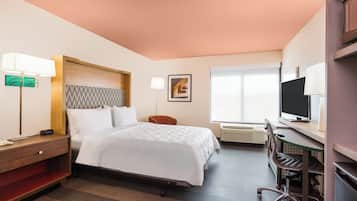Habitación de hotel con cama, mesita de noche, lámpara, silla, escritorio, televisor y aire acondicionado.