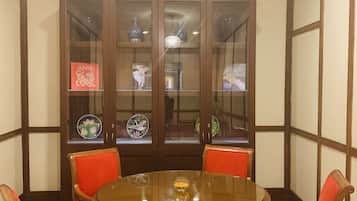 A dining area with a round table surrounded by six chairs with red cushions, a wooden cabinet with glass doors displaying various items, and a ceiling with recessed lighting.